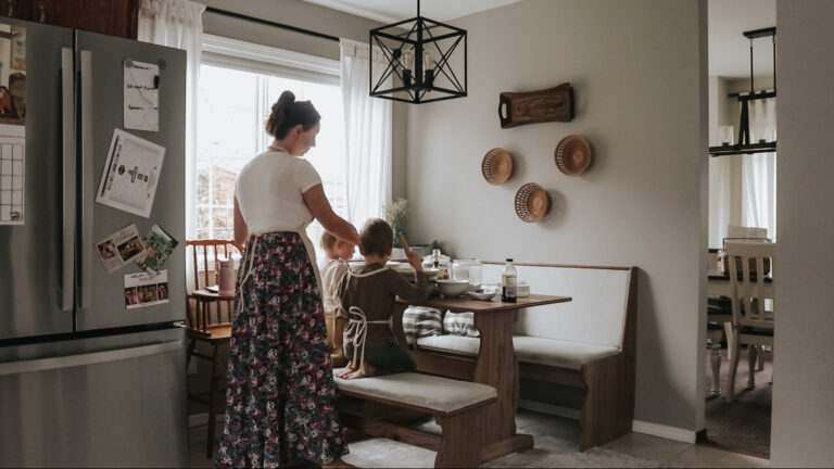 Christian homemaker stands at kitchen table where her children are sitting and instructs them on how to make muffins